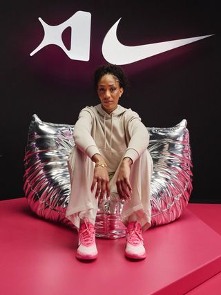 A'ja Wilson wearing a tan sweatshirt and sweatpant set and her new hot pink A'One Nike signature sneakers while sitting on a silver lounge chair.