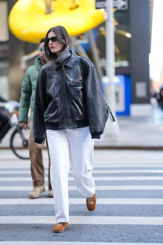 A woman wears a leather jacket, white jeans, and tan suede sneakers.