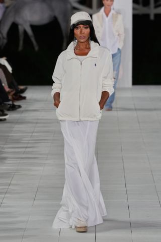 Naomi Campbell wearing a baseball cap with a track jacket and white maxi dress at the Ralph Lauren show.