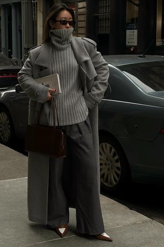 A woman standing on the sidewalk wearing a gray wool trench coat over a gray ribbed turtleneck, gray trousers, brown pointed heels, and a brown bowler bag.