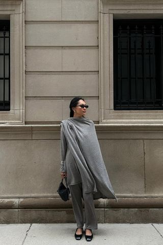 A woman standing on the sidewalk wearing black sunglasses with a gray cape over a knit top and matching knit pants, a chunky silver bangle, black ballet flats, and a black handbag.