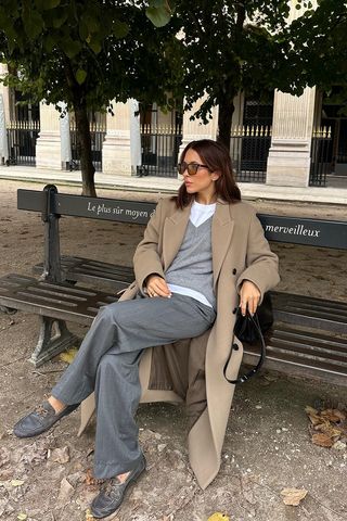 A woman sitting on a park bench in Paris wearing brown sunglasses, a brown long coat, a white t-shirt, a gray v-neck sweater, gray trousers, a black tote bag, and gray boat shoes.