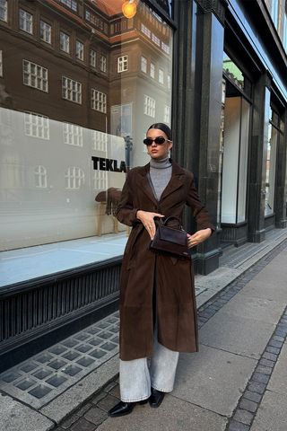 A woman standing on the sidewalk in Copenhagen wearing brown sunglasses with a gray turtleneck sweater, gray stone-wash jeans, brown ankle boots, a brown crossbody bag, and a brown suede trench coat.