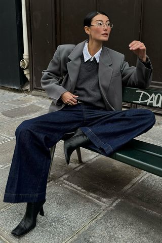 A woman sitting on a bench outside wearing a gray blazer over a white button-down shirt, a gray v-neck sweater, dark wash wide-leg jeans, and black ankle boots.