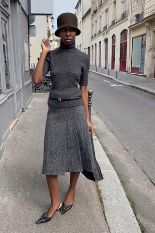 A woman standing on the sidewalk wearing a brown hat, a gray turtleneck sweater, a black skinny belt, a gray pleated midi skirt, black mules, and a black handbag.