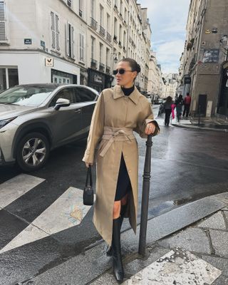 Fashion influencer @cocoschiffer in Paris wearing an anti-trend outfit styled with a trench, a turtleneck, a black skirt, and knee-high boots.