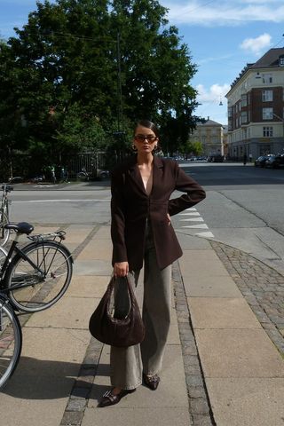 dark brown outfit idea shown in a photo of a woman standing on the sidewalk in Copenhagen wearing brown sunglasses, a brown nipped-waist blazer, brown jeans, brown Ganni ballet flats, and a brown woven hobo handbag