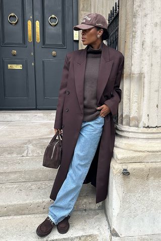 dark brown outfit idea shown in a photo of a woman standing on the stairs in front of a brownstone in London wearing a dark brown baseball cap, long brown coat, a turtleneck sweater, straight-leg jeans, suede loafers, and a suede bowler bag