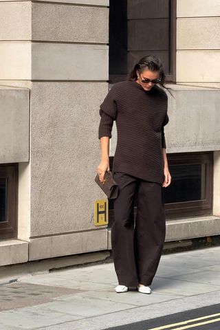 dark brown outfit idea shown in a photo of a woman standing on the sidewalk in London wearing brown sunglasses with a brown asymmetrical sweater, brown barrel-leg trousers, white mules, and a brown suede clutch bag