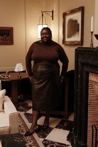 dark brown outfit idea shown in a photo of a woman standing inside a hotel lobby near a fireplace wearing a dark brown asymmetrical sweater with a dark brown leather midi skirt and brown pointed mules