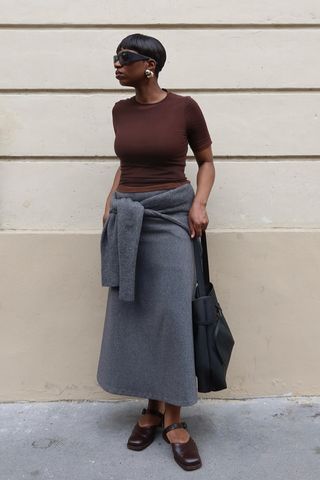 dark brown outfit idea shown in a photo of a woman standing on the sidewalk in Paris wearing oversized black sunglasses, gold earrings, a brown t-shirt, a gray midi skirt with a gray cardigan tied around the waist styled with a black tote bag and brown Mary Jane flats