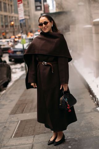 dark brown outfit idea shown in a photo of a woman standing on the sidewalk in New York wearing a black headband, brown sunglasses, silver earrings, a brown scarf coat, a brown belt, a black bowler bag, and brown pointed flats