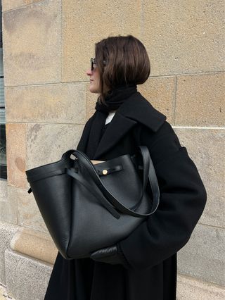 Eliza Huber testing out the Tory Burch Romy Tote Bag in black with a long black Nanushka coat, jeans, and a scarf.