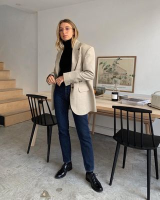 British influencer Brittany Bathgate poses in front of a stylish dining table wearing wire-rimmed optical eyeglasses an oversize beige blazer, black turtleneck, dark-wash skinny jeans, and black lace-up oxfords