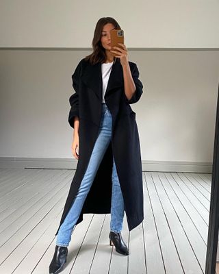 British influencer Marianne Smyth poses for a mirror selfie wearing a long black coat, white T-shirt, skinny jeans, and square-toe kitten heel boots