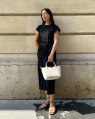 A woman wears a black dress, long champ bag, and beige sandals.