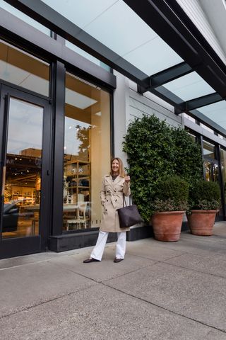 A Nordstrom stylist wearing a tan trench coat, white jeans, and ballet flats.