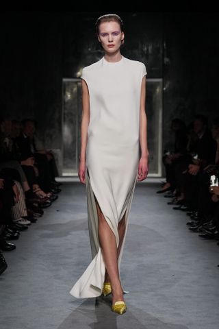 A model wearing a white gown with yellow satin kitten heels walking in Haider Ackermann's first Tom Ford runway show during Paris Fashion Week.