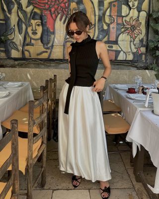 fashion influencer Debora Rosa poses at the La Colombe d'Or restaurant in Provence wearing black oval sunglasses, a black ribbed tank top, black skinny scarf, a satin white bubble hem skirt, and strappy black sandals