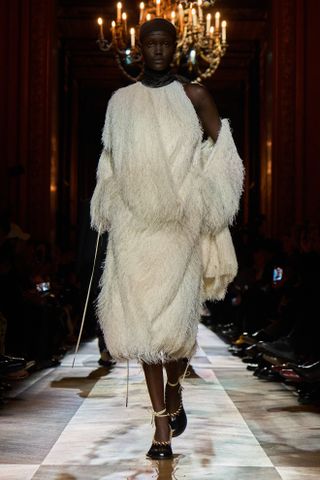 A model wearing a white fringe coat and dress while walking at the F/W 25 Dries Van Noten show during Paris Fashion Week.