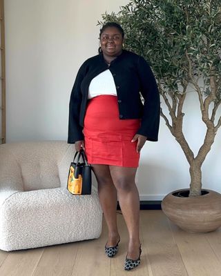 British plus size female fashion influencer poses next to a boucle chair and olive tree in a living room wearing a black cardigan sweater, white top, red mini skirt, and Gucci crystal kitten heel slingback shoes