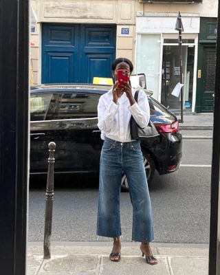 European style influencer Sylvie Mus poses for a mirror selfie on the streets of Paris wearing a white button-down shirt, cropped wide-leg jeans, and black kitten-heel mules.