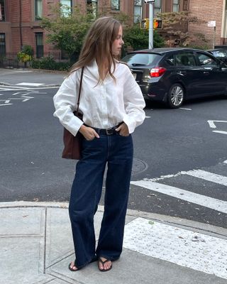 Danish fashion creative Clara Dyrhauge poses on a NYC street wearing a white linen button-down shirt, a brown tote bag, black belt, dark-wash baggy jeans, and black flip-flop sandals.