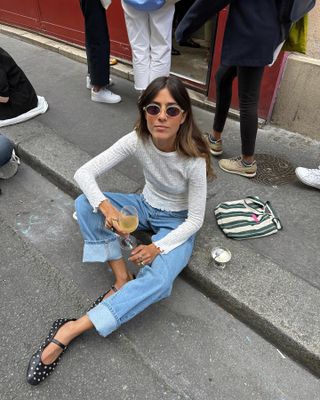 Portugese style influencer Vicky Montanari sits on a sidewalk holding a glass of wine and wearing a pair of metal-rim sunglasses, a sheer ribbed white T-shirt, cuffed jeans, and studded Mary Jane flats.