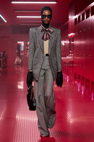 A model walking in the F/W 25 Valentino show during Paris Fashion Week wearing a black balaclava, gray suit, pussy bow blouse, and fur gloves.