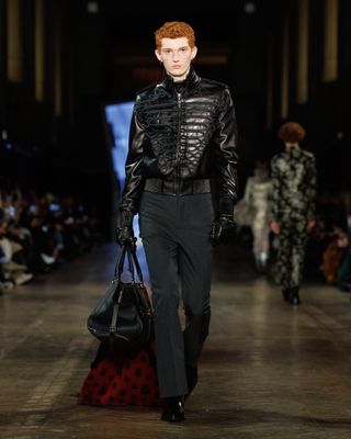 A male model wearing a leather jacket and trousers at the F/W 25 Alexander McQueen show carrying a black weekender bag adorned with a red-and-black skull scarf.