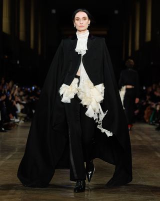 A model wearing a while silk blouse, black cape coat, and black trousers while walking at the F/W 25 Alexander McQueen show.