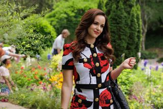 Melissa Fumero playing Birdie in Grosse Pointe Garden Society. She is standing in a garden wearing a rose-print top and skirt set.