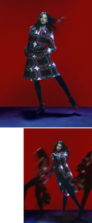 Simone Ashley poses mid-movement in front of a red backdrop wearing a geometric-print red, white, and blue coat with black tights and red heels.