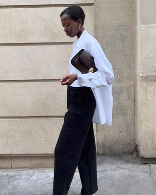 European fashion influencer Sylvie Mus wearing a polished white button-down shirt, a chic brown clutch bag, and black pants.