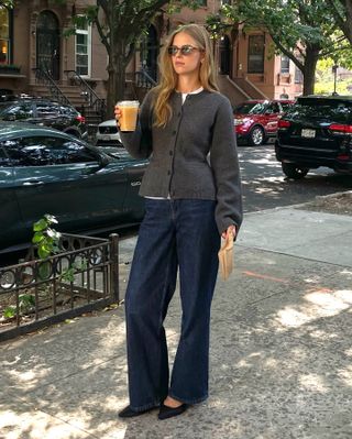 Danish fashion creative Clara Dyrhauge poses on a NYC sidewalk holding a coffee wearing brown rectangular sunglasses, a gray waisted cardigan layered over a white T-shirt, and black kitten heels